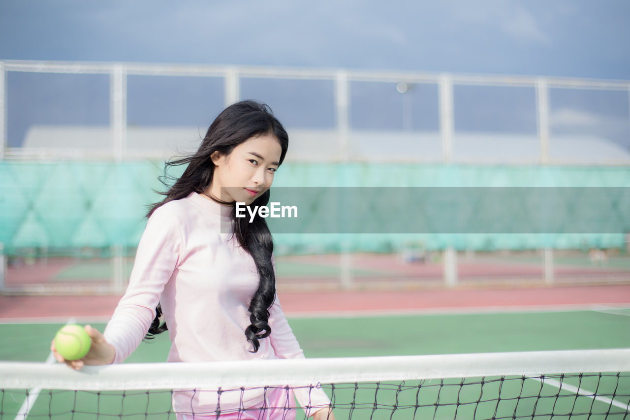 Portrait of young woman holding tennis ball while standing against net on court