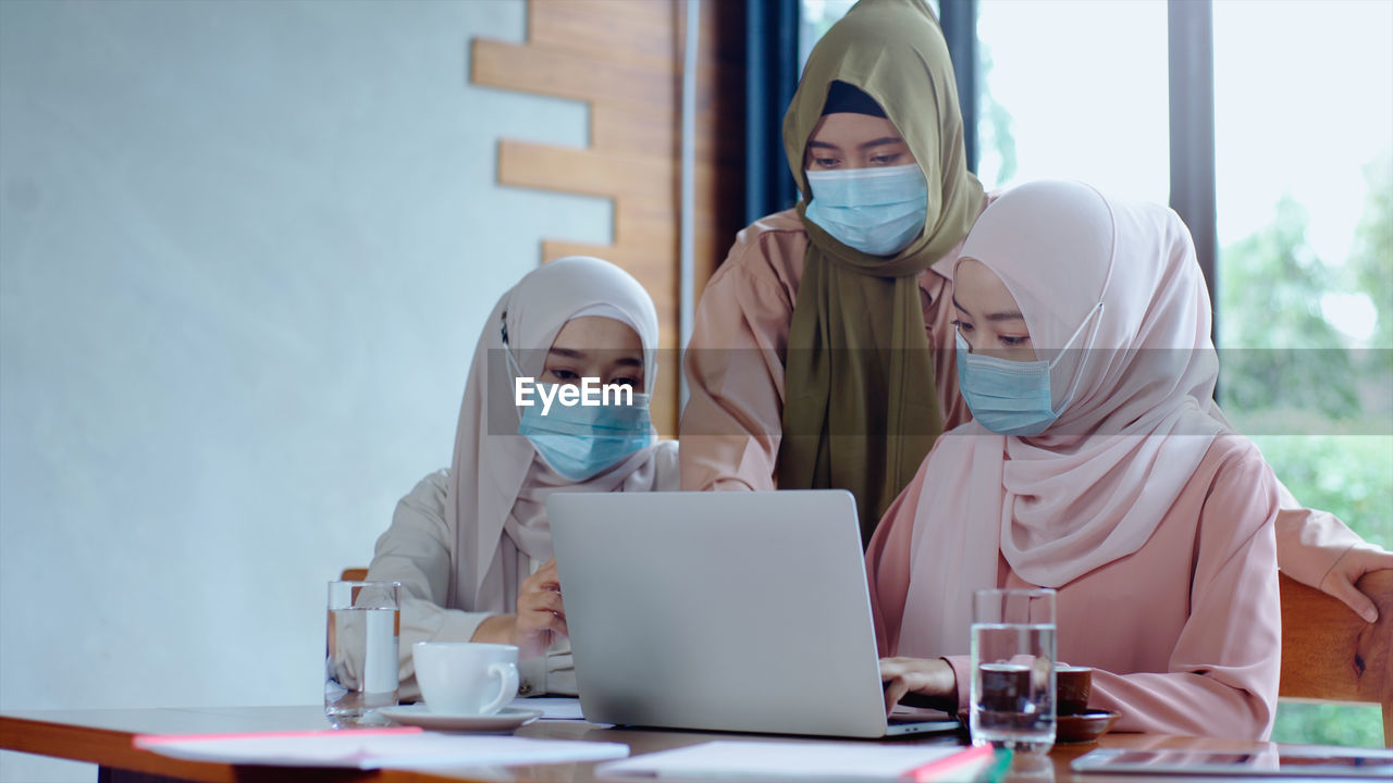 Businesswomen in hijab wearing mask looking at laptop in office