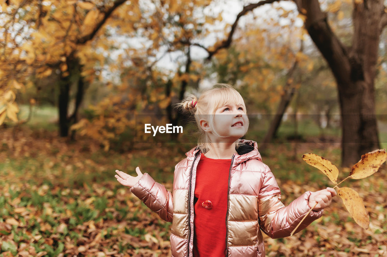 Happy little blonde girl plays with yellow autumn leaves in garden. kid throwing up leaves in park