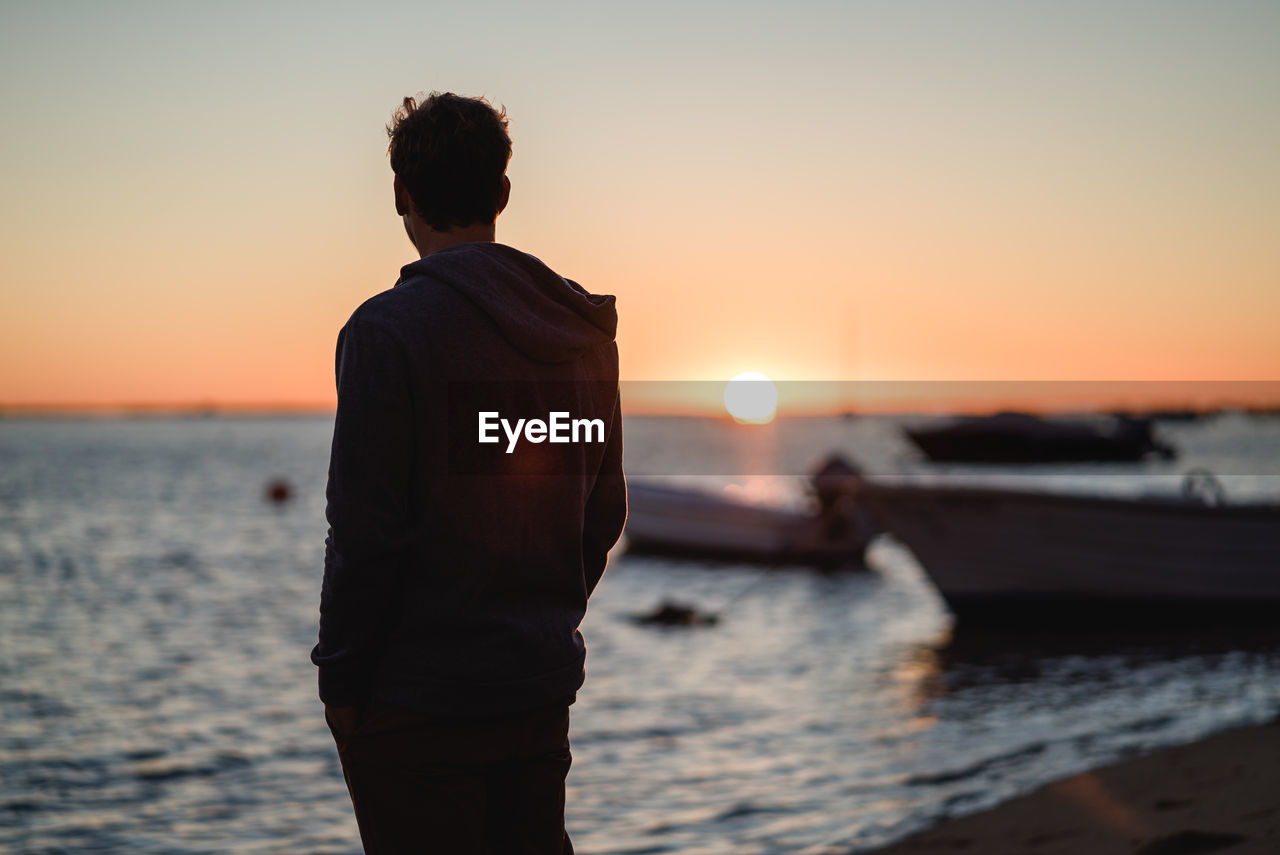 Rear view of man standing at beach against sky during sunset