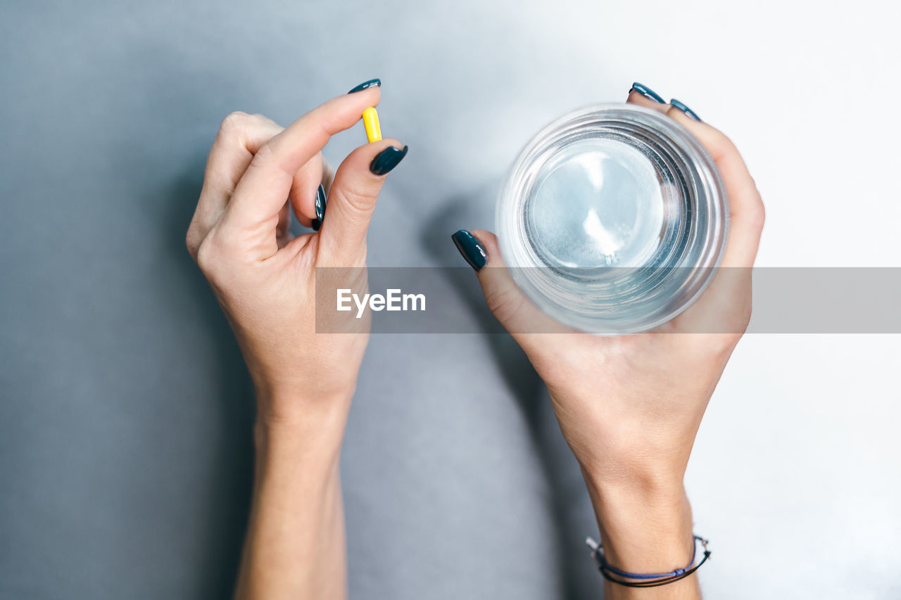 Hands of caucasian girl with yellow pill in left palm and glass of water in right hand. 