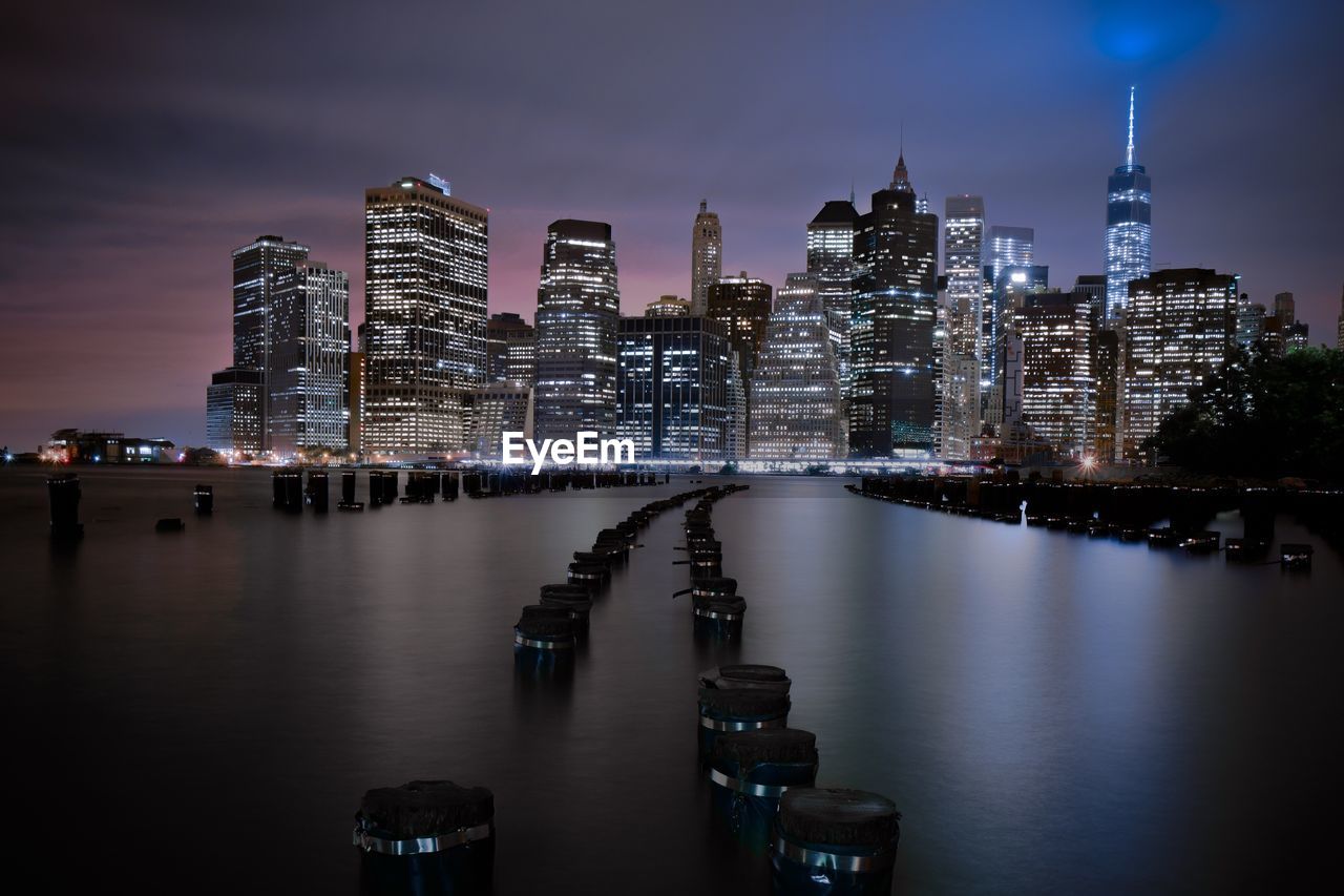 Wooden posts amidst river against cityscape at night