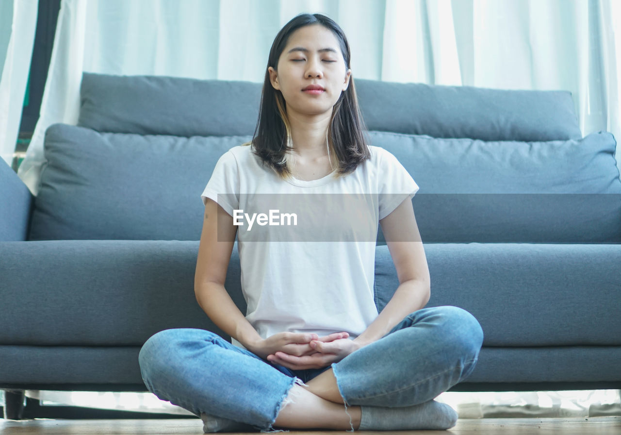 YOUNG WOMAN LOOKING AWAY WHILE SITTING ON SOFA AT HOME