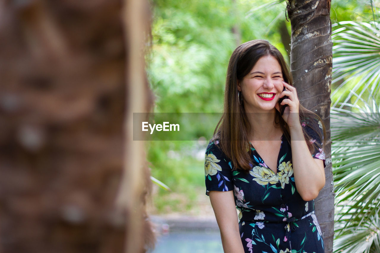 Beautiful young woman using phone while standing against tree trunk