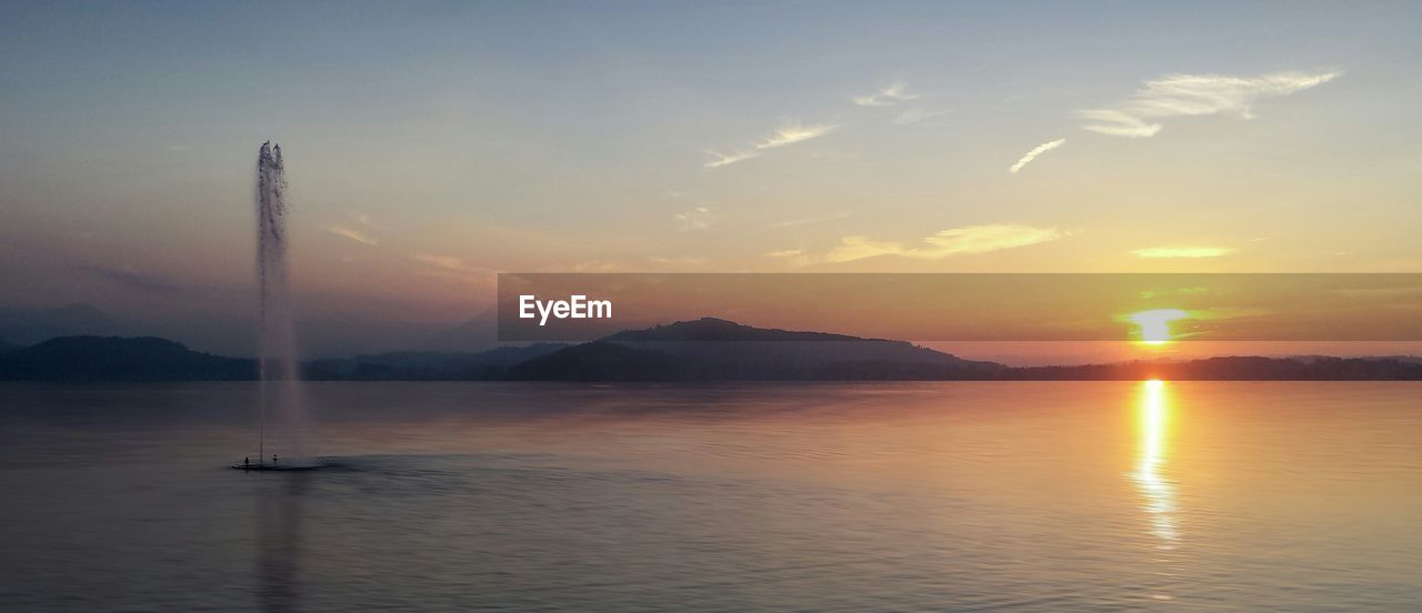 Fountain in lake in front of mountains during sunset