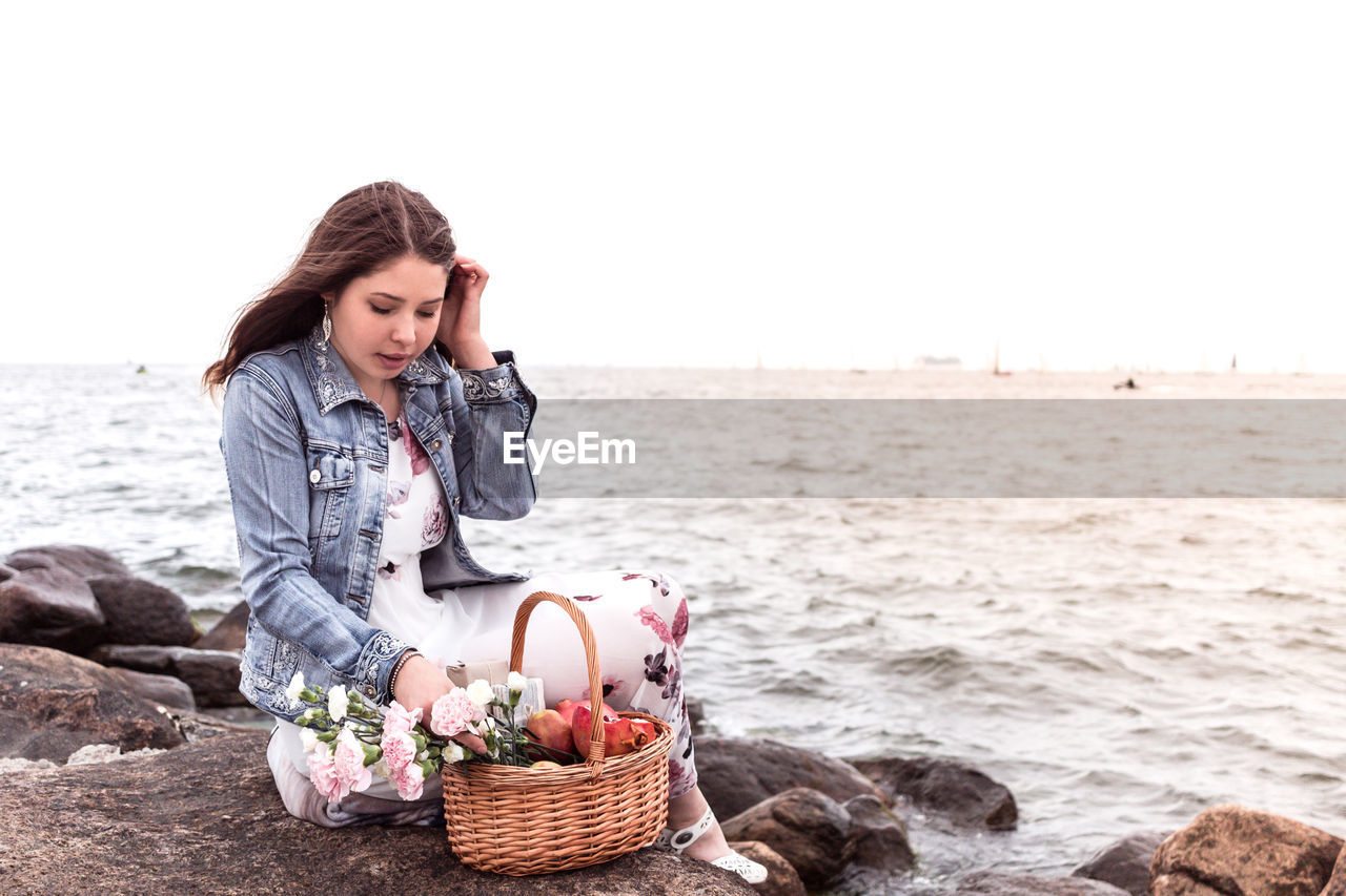 Young woman with wicker basket sitting on rock at beach against clear sky