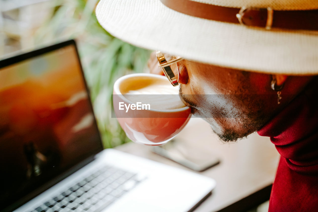 From above cropped unrecognizable trendy african american male entrepreneur sitting in cafe and enjoying fresh coffee during break at remote work