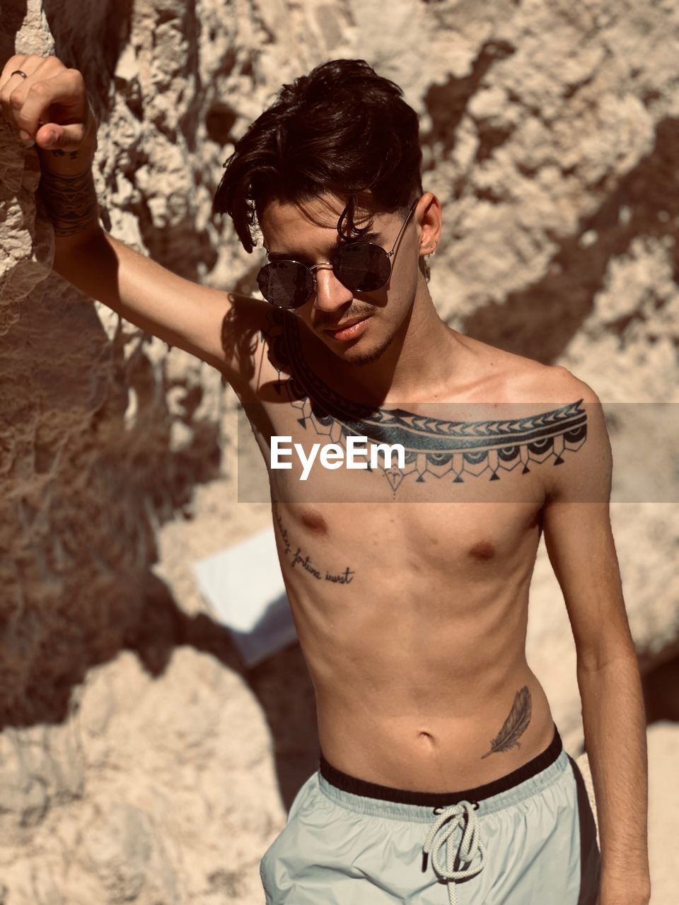 Shirtless man with tattoo wearing sunglasses leaning on rock formation