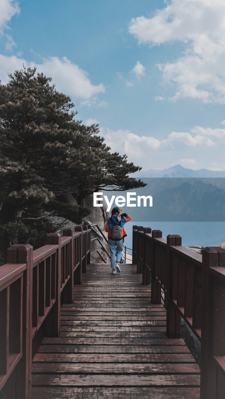 Rear view of woman walking on footbridge against sky