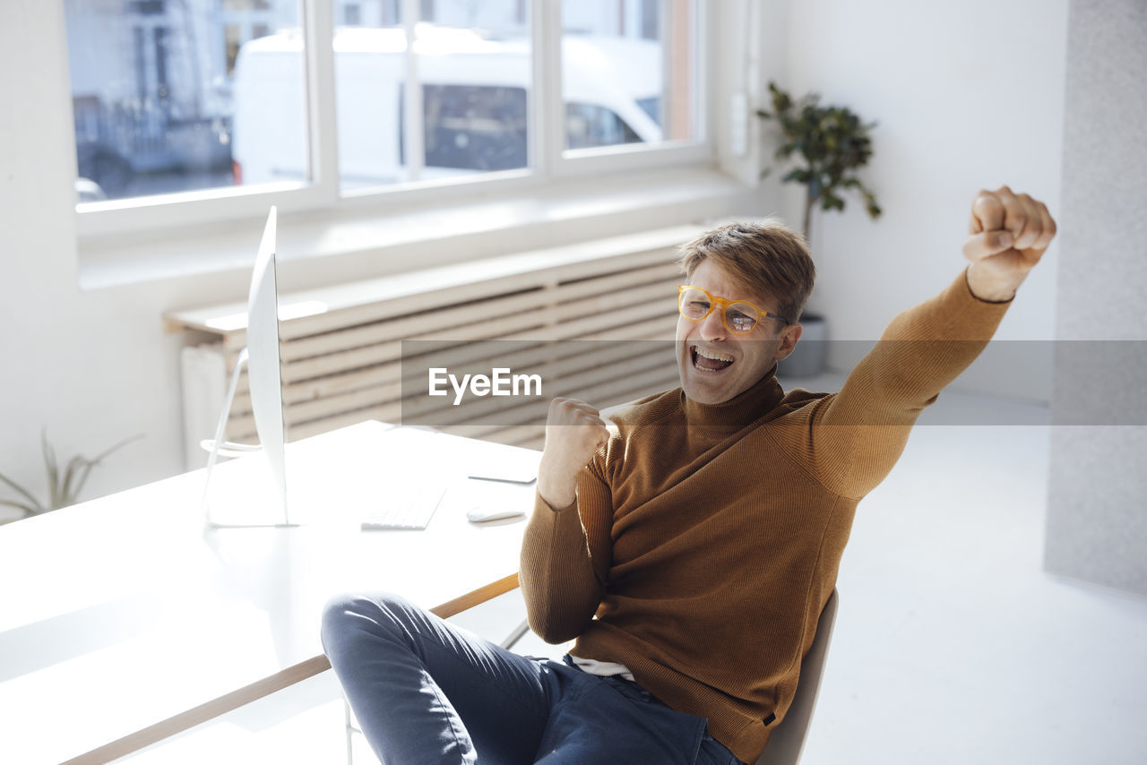 Happy businessman gesturing fist sitting at desk in office