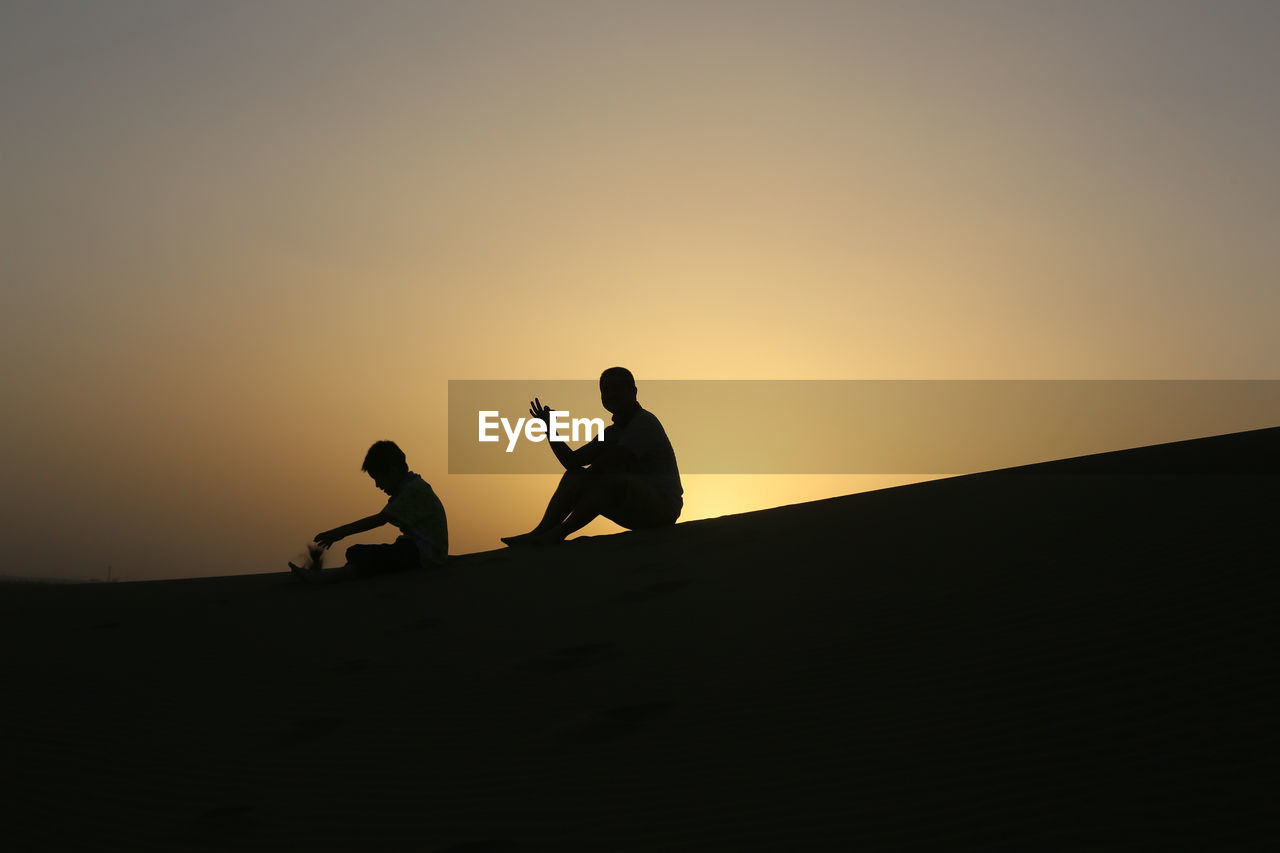 Silhouette of father and son sitting on sand