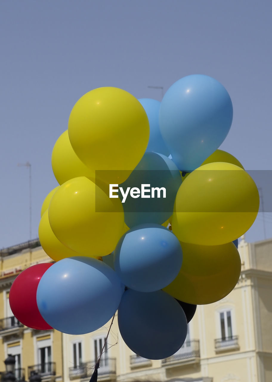 balloon, toy, helium balloon, helium, blue, multi colored, celebration, event, no people, nature, yellow, bunch, anniversary, architecture, large group of objects, sky, party, birthday, mid-air, building exterior, clear sky, city, day, built structure, outdoors, decoration, flying, group of objects