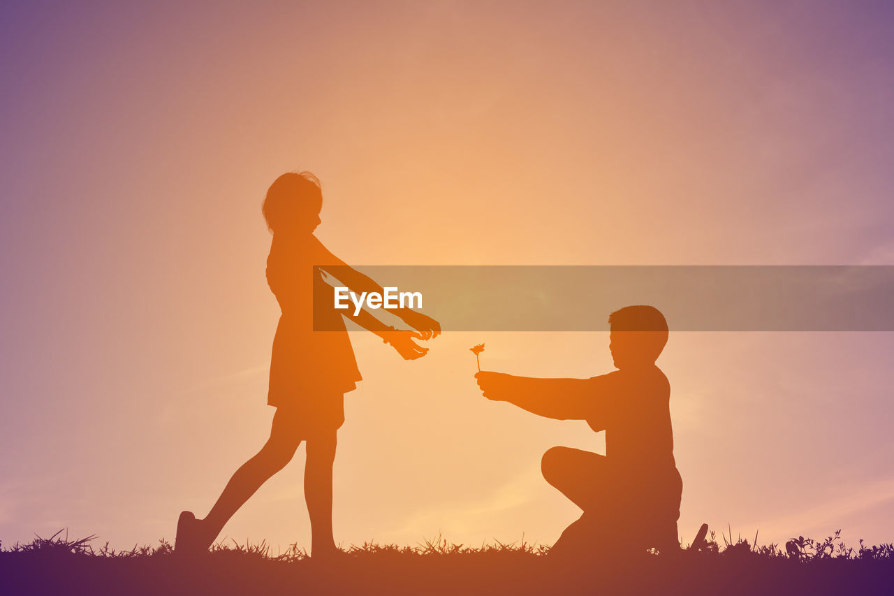 Low angle view of teenage boy offering flower to girl on field against sky during sunset