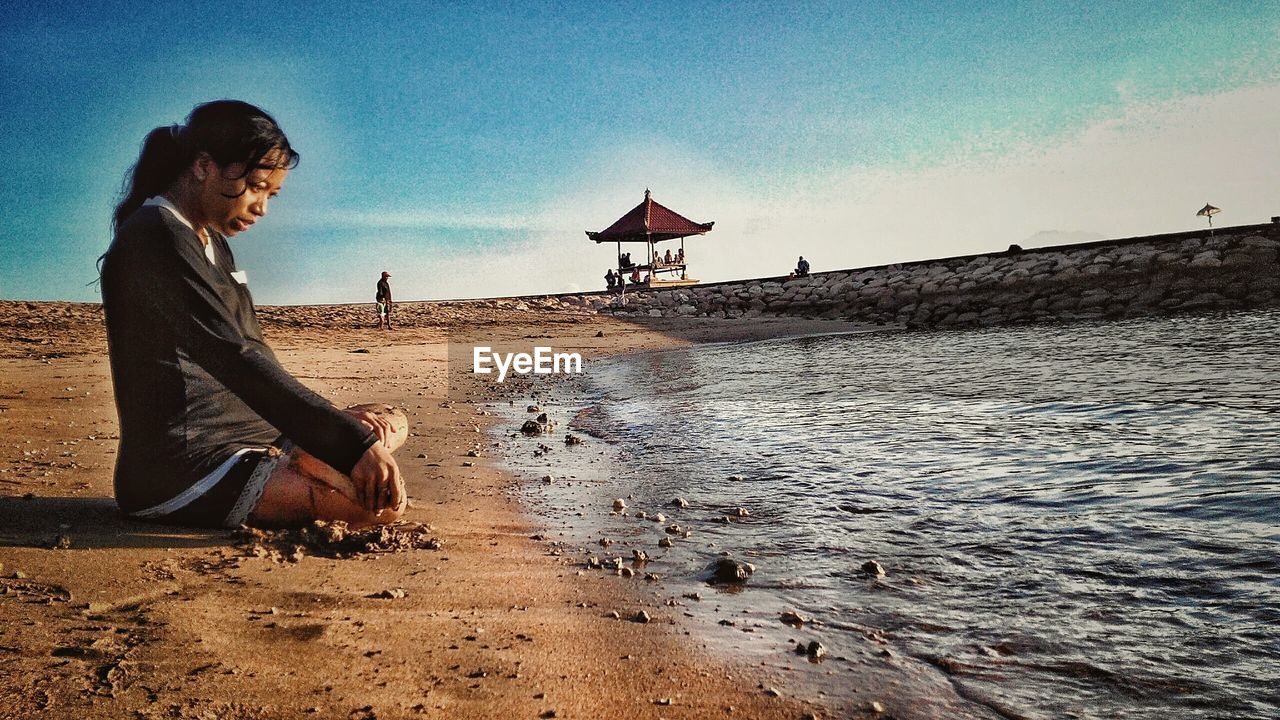 SIDE VIEW OF YOUNG WOMAN SITTING ON BEACH AGAINST SKY