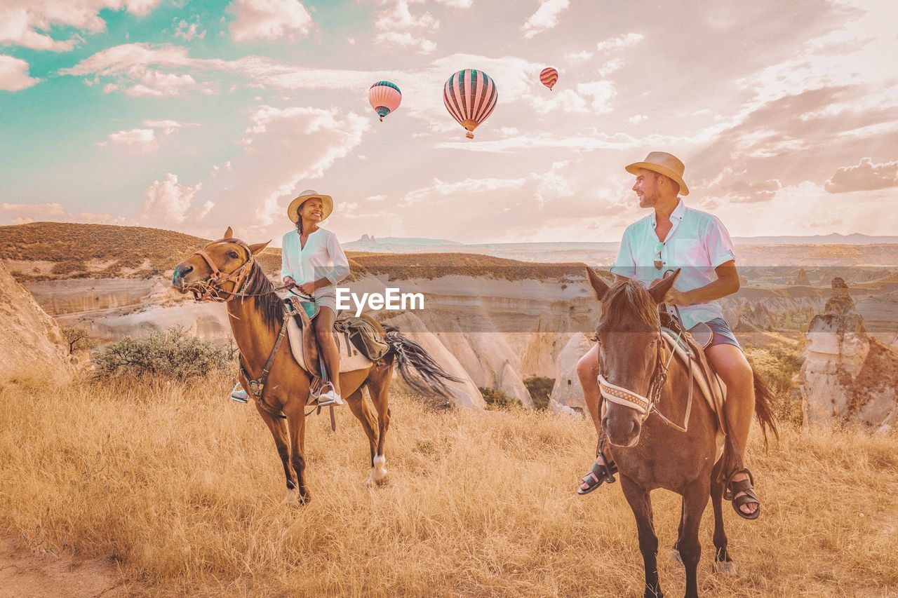 Couple sitting on horses against hot air balloons flying in mid-air