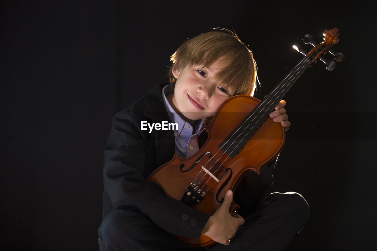 Boy holding violin while sitting against black background