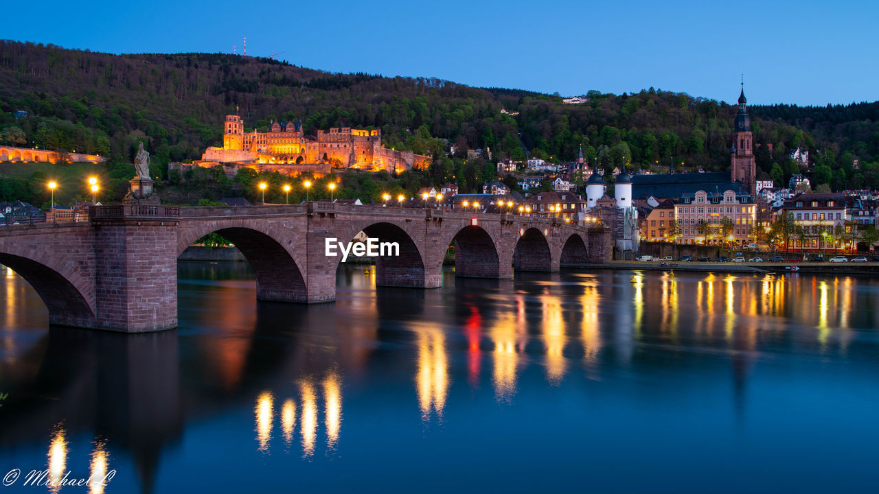 ILLUMINATED BRIDGE OVER RIVER BY BUILDINGS AGAINST SKY