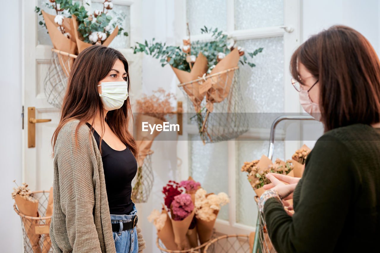 Female customer in protective mask consulting with saleswoman working in store with different bunches of flowers wrapped in zero waste paper package