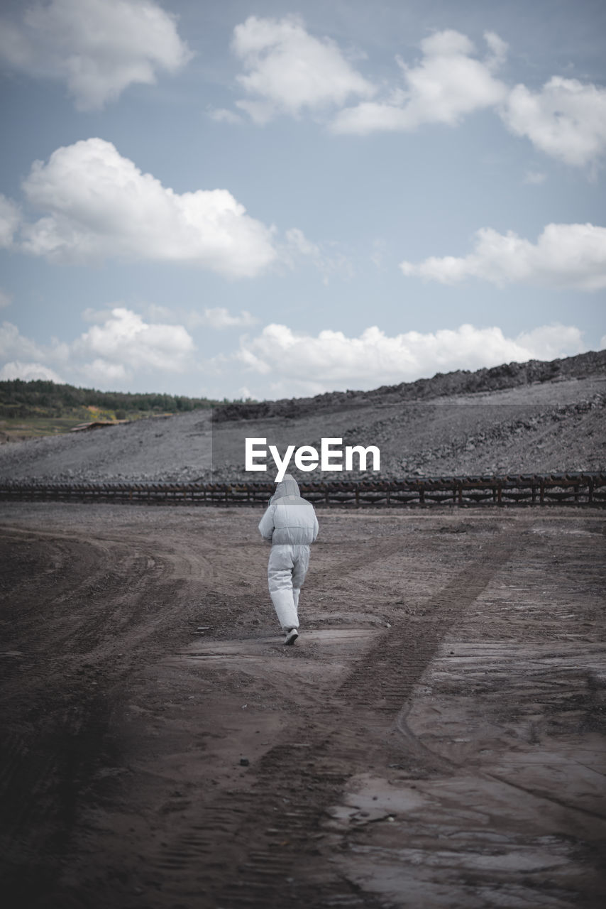 Rear view of worker in protective clothing walking on field at mining industry against sky