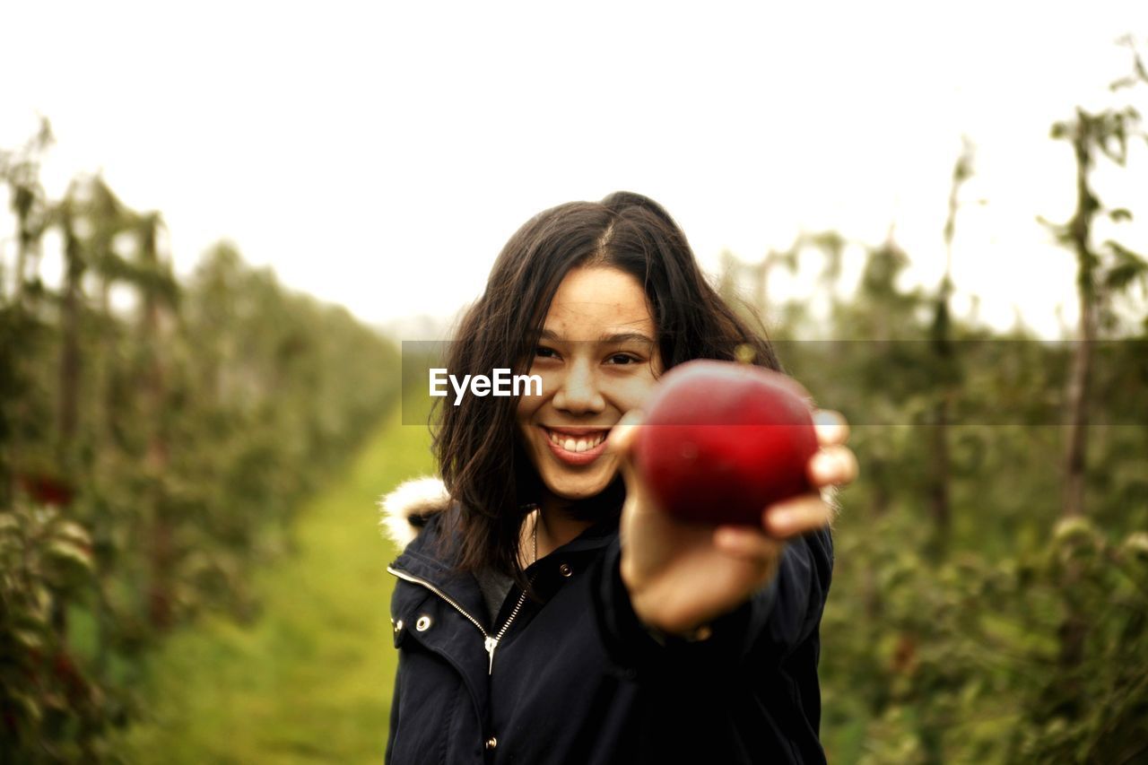 Portrait of smiling young woman holding apple against sky