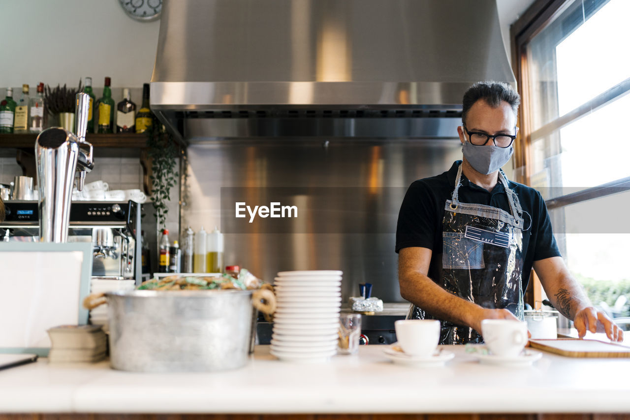 Male barista wearing protective face mask while working in kitchen of coffee shop during covid-19