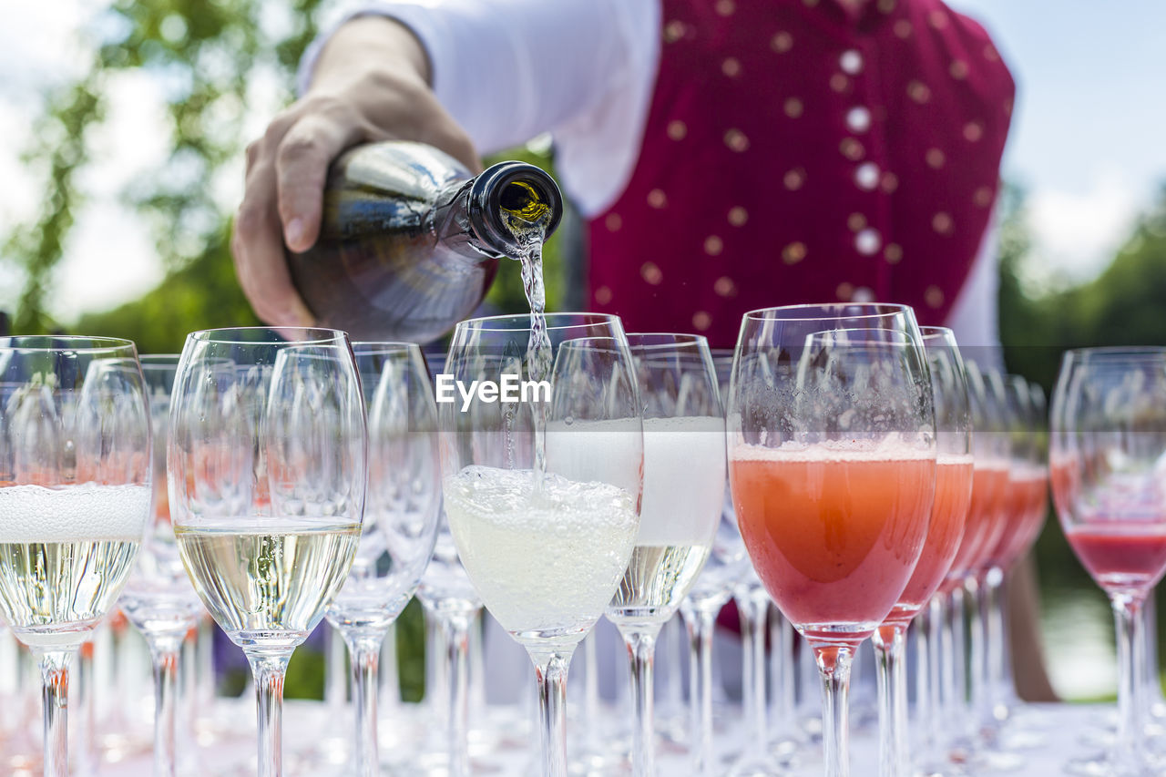 Midsection of bartender pouring aperitif in champagne flutes on table during outdoor party