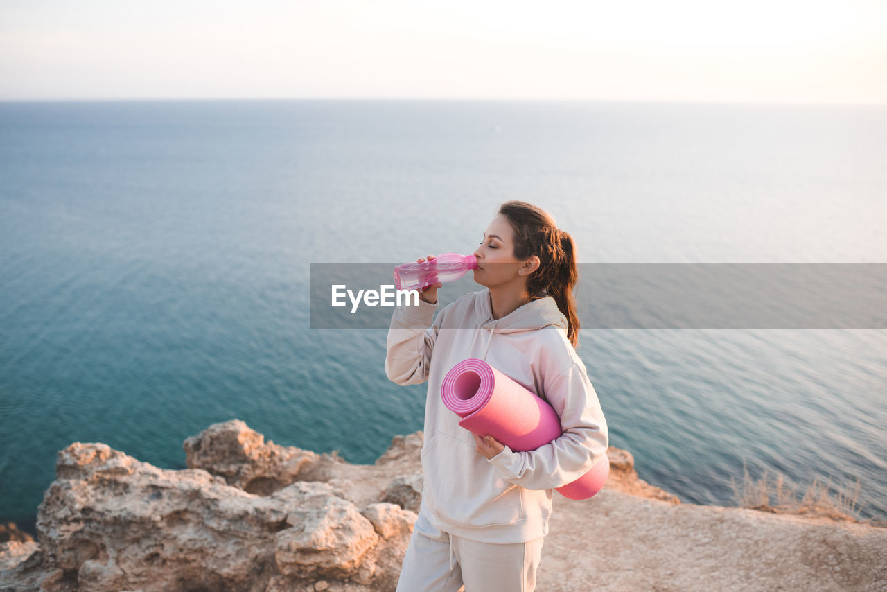 Pretty woman drinking water from pink bottle wear spotswear hoodie and pants hold yoga mat over sea