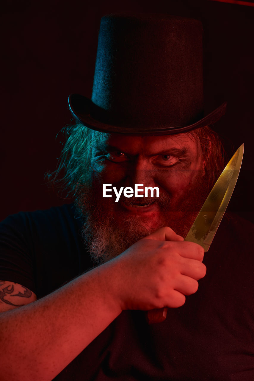 hat, adult, one person, fedora, cowboy hat, men, knife, portrait, clothing, facial hair, weapon, person, melee weapon, fashion accessory, evil, red, beard, dark, holding, costume hat, aggression, human face, indoors, studio shot, mature adult, emotion, sign, darkness, headshot, spooky, anger, looking at camera, activity, social issues, kitchen knife