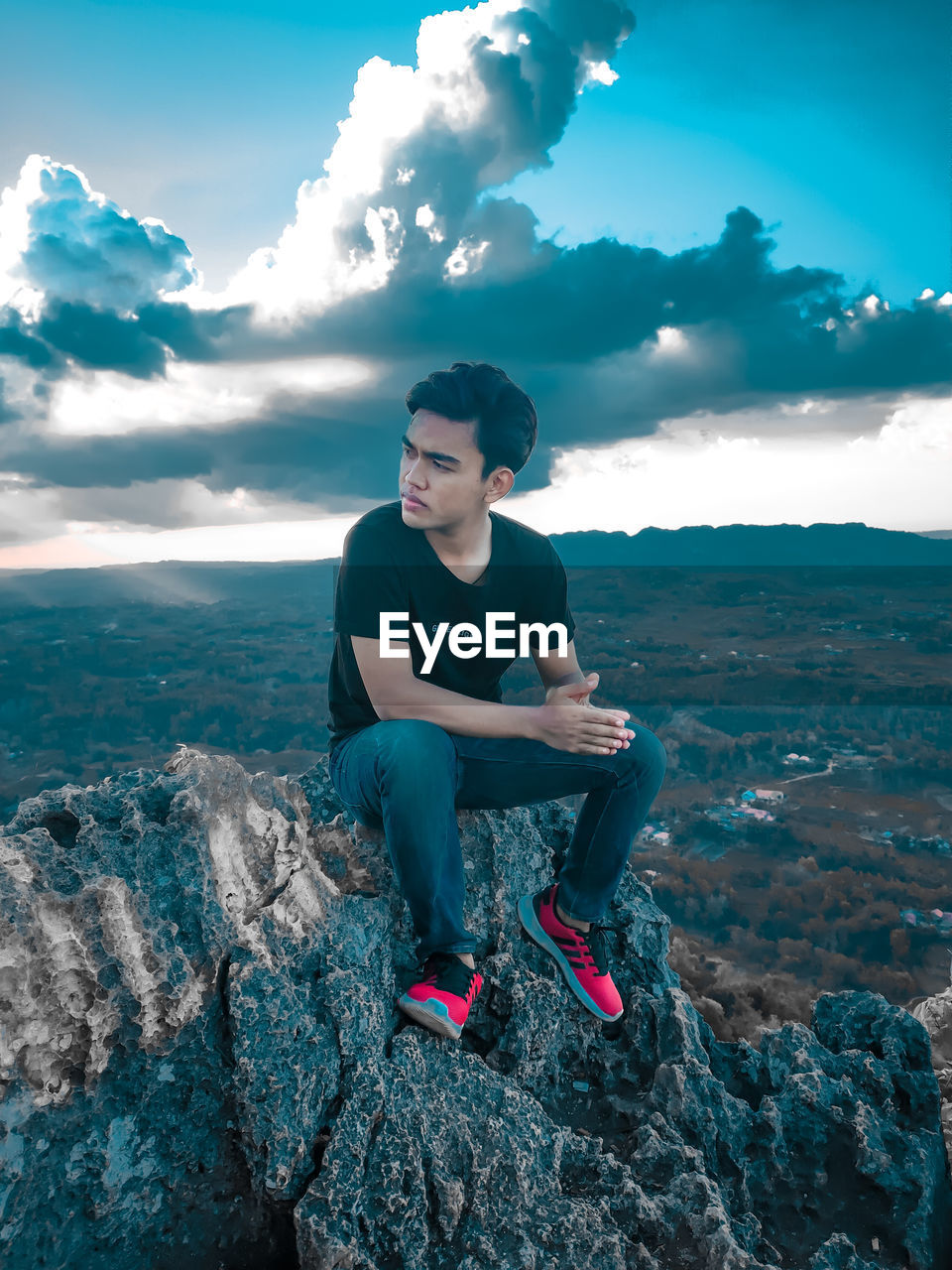 YOUNG MAN SITTING ON ROCK AGAINST SKY
