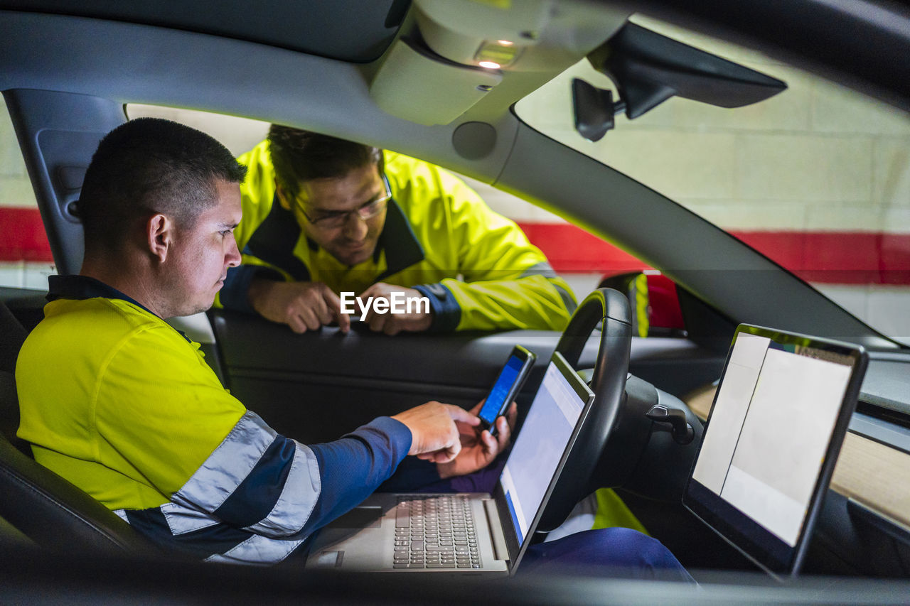 Male programmer showing mobile phone to colleague while sitting in electric car