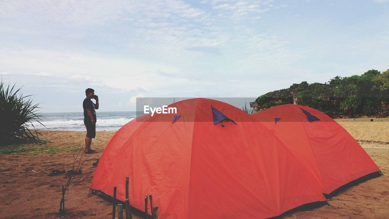 Man talking on phone while standing by tent at beach against sky