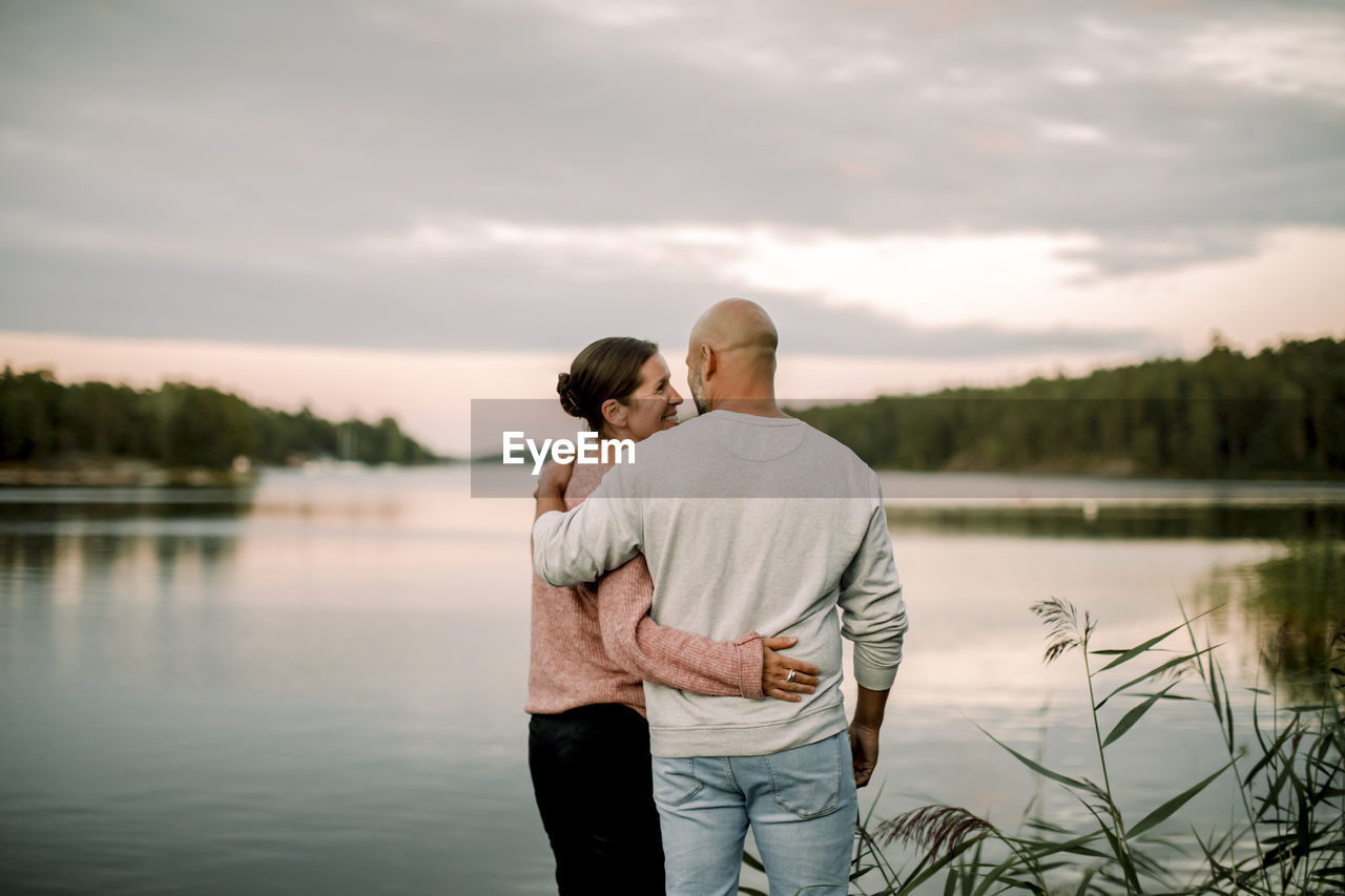 Rear view of smiling couple with arm around standing against lake during sunset