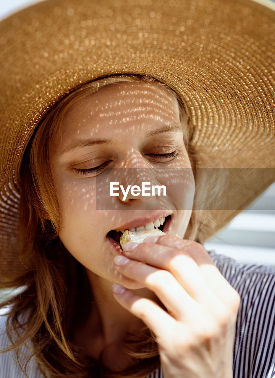 Woman in hat eating grilled vegetables outdoors, eyes closed. light and shadow pattern on the face