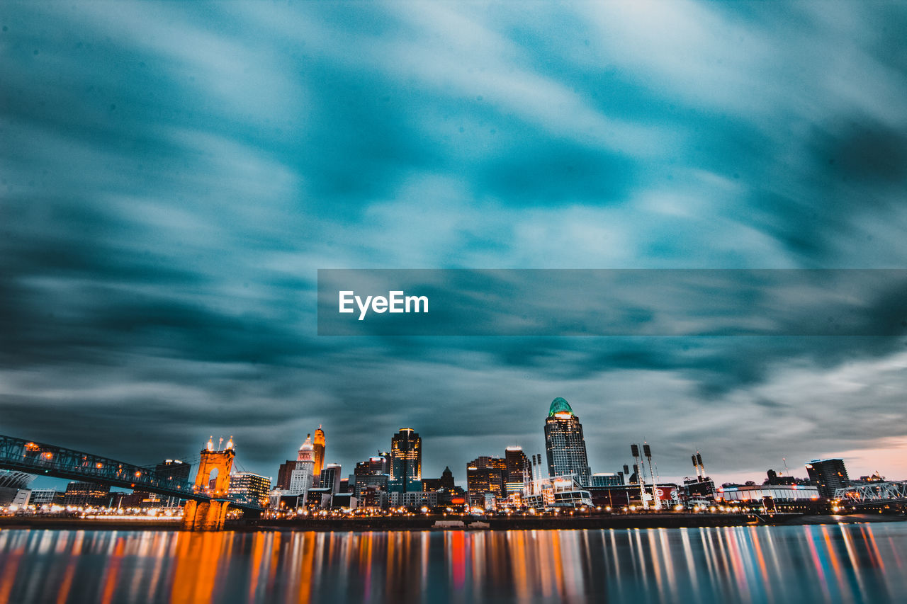 Illuminated city at waterfront against cloudy sky