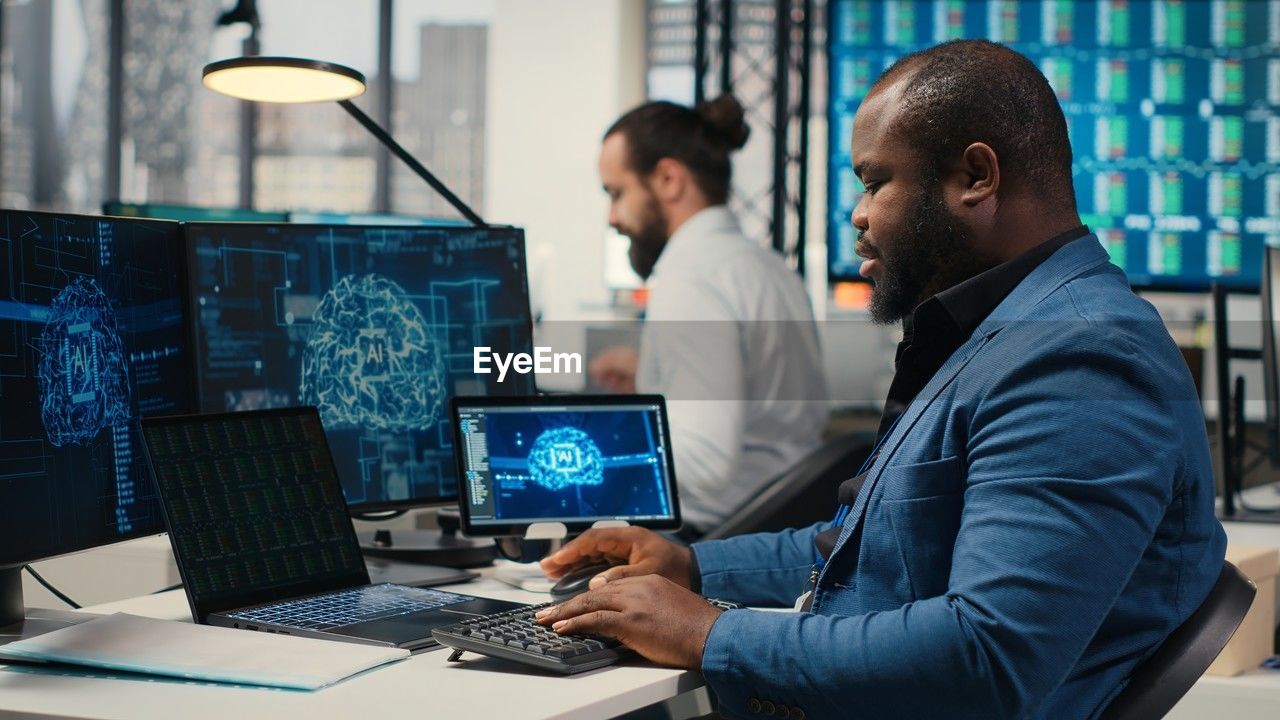 technology, computer, adult, men, communication, business, working, occupation, wireless technology, sitting, office, two people, indoors, cooperation, computer equipment, teamwork, computer monitor, table, laptop, businessman, business finance and industry, using computer, furniture, desk, person, young adult, internet, computer network, corporate business, coworker, looking, planning, waist up, businesswear, discussion, using laptop, concentration, expertise, side view, professional occupation, desktop pc