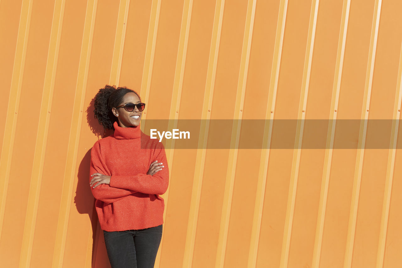 Happy woman wearing sunglasses in front of orange wall on sunny day