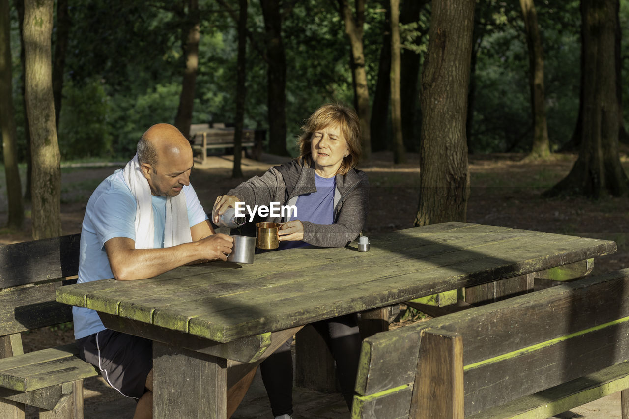 Senior couple sitting in picnic table in forest drkinking water from bottles in metal camping cups