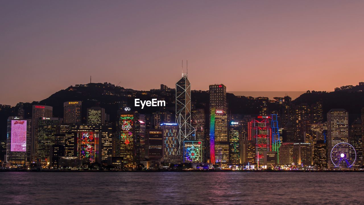 Illuminated modern buildings by river against clear sky at dusk