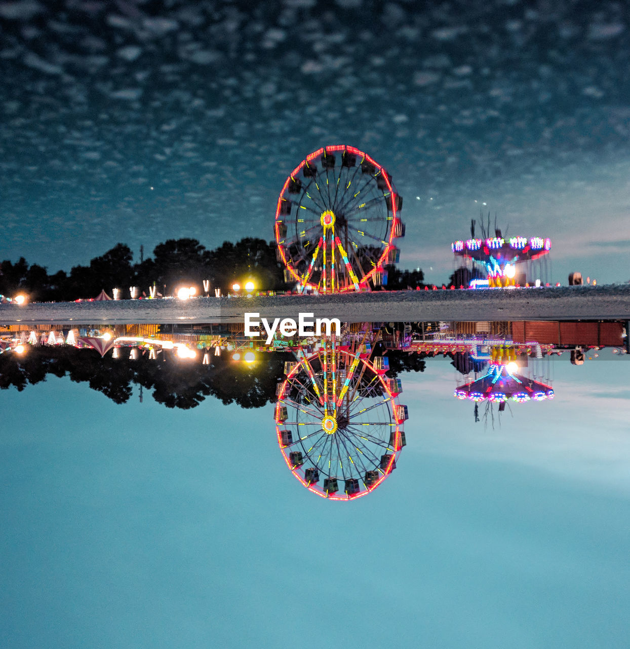 Reflection of ferris wheel on calm water against cloudy sky at night