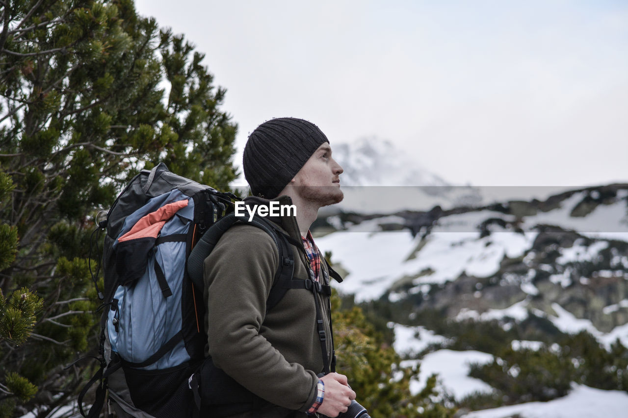 Side view of hiker with backpack while standing on snow covered mountain against sky