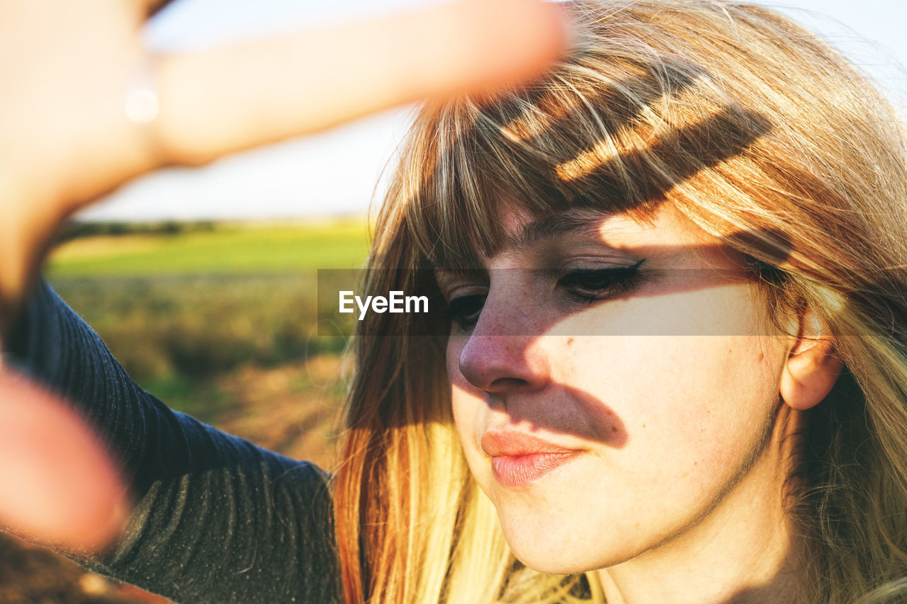 Close-up of young woman shielding eyes while standing at field against sky on sunny day