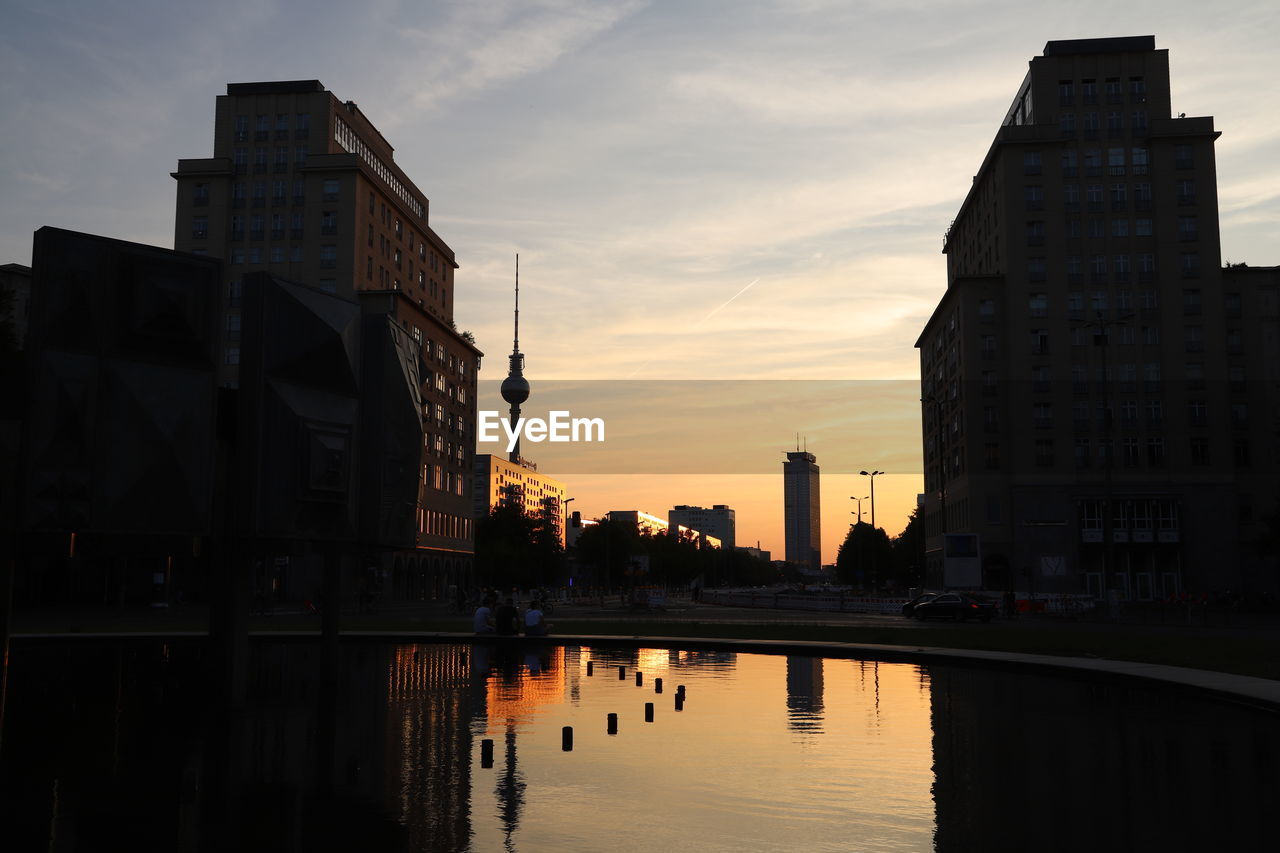 Silhouette buildings by lake against sky at sunset