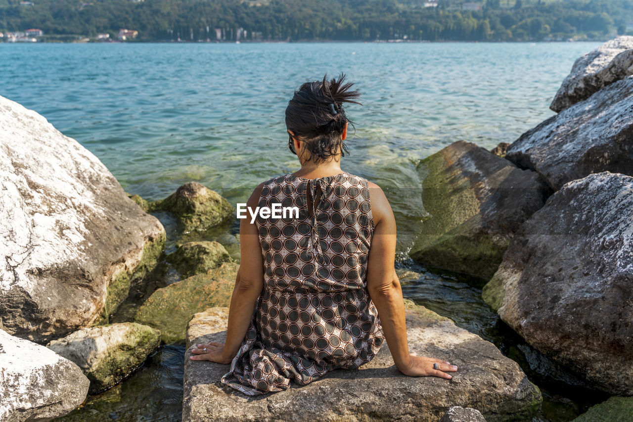 Rear view of woman looking at lake while sitting on rock