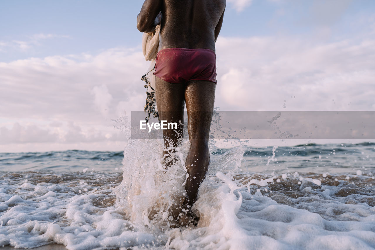 Shirtless fisherman walking with fishing net towards sea against sky