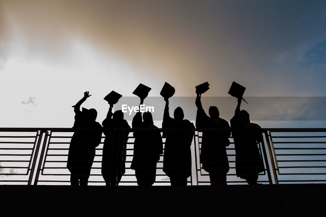 Silhouette people throwing cap while standing by railing against sky