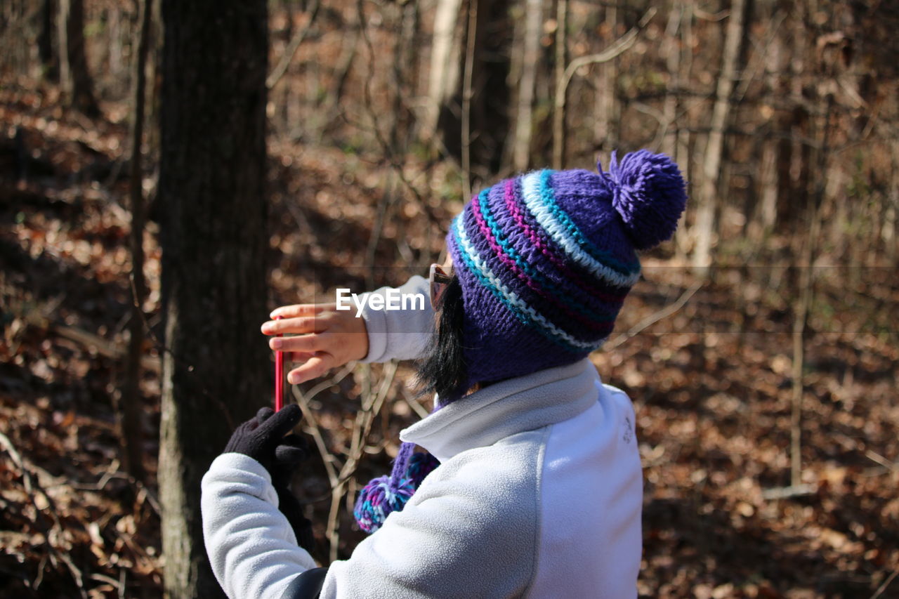 Rear view of woman wearing knit hat using phone at forest