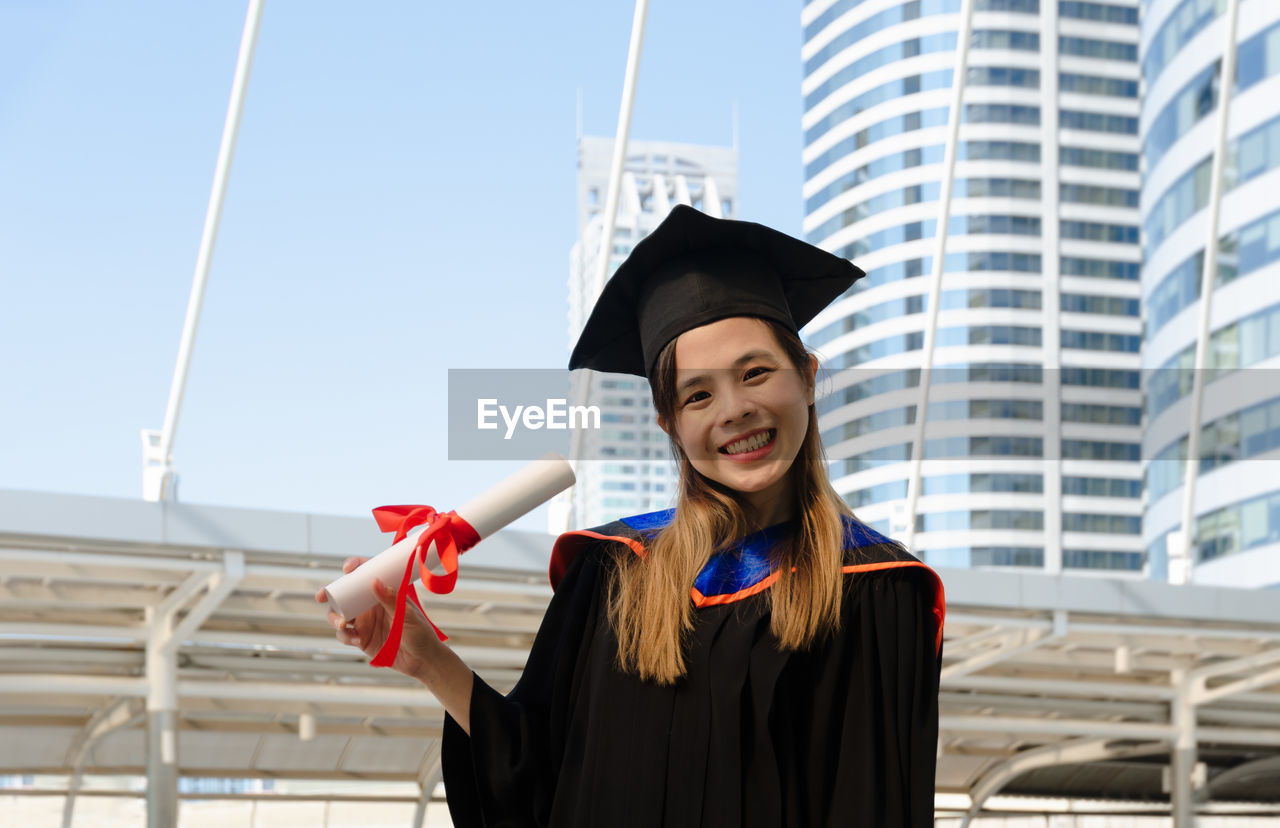 PORTRAIT OF SMILING YOUNG WOMAN STANDING AGAINST BUILDING