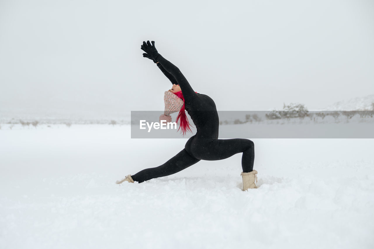 Side view of flexible female practicing yoga in anjaneyasana while standing in snowy field in winter