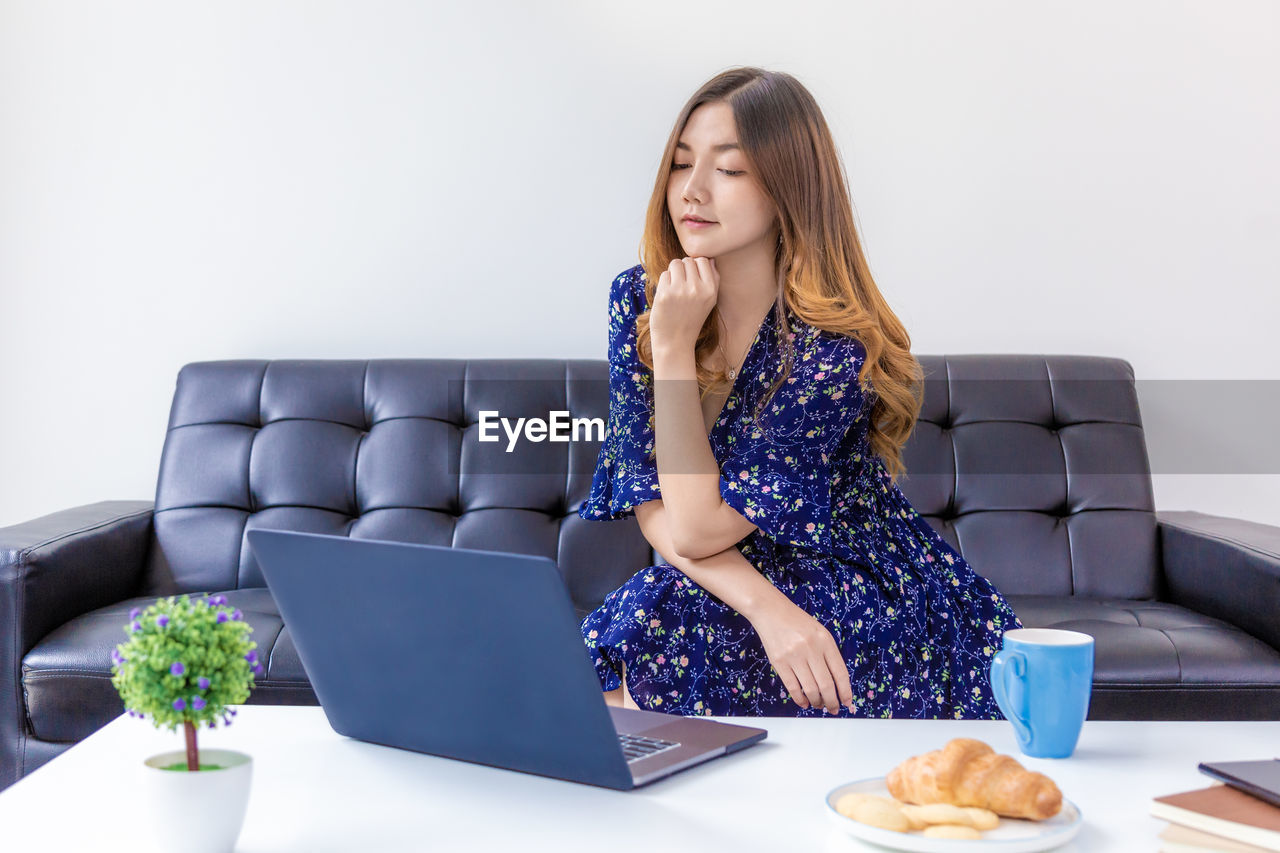 YOUNG WOMAN USING MOBILE PHONE WHILE SITTING ON SOFA AT HOME