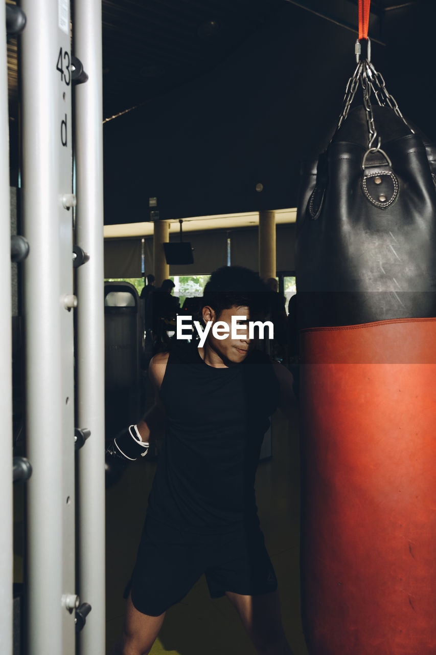 Young focused asian man training boxing performing punches while exercising with heavy punching bag in a modern gym